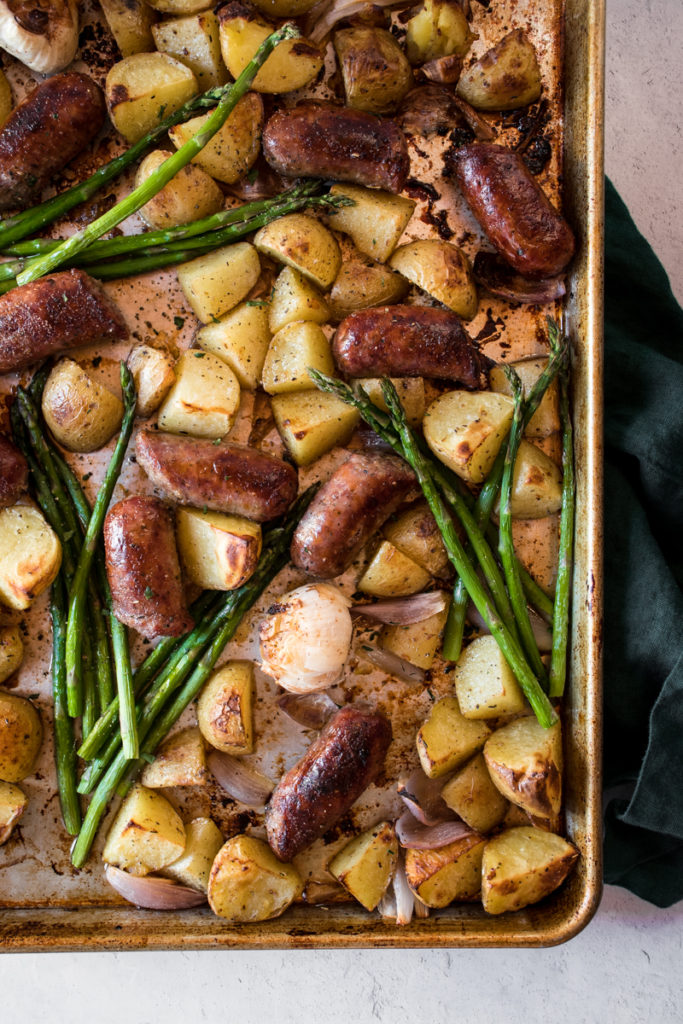 Roasted italian sausages and crispy potatoes with tender asparagus arranged on a sheet pan with roasted garlic cloves.
