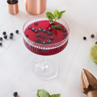 deep purple hued cocktail garnished with blueberries and fresh mint served in a coupe glass