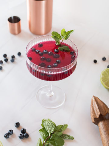 deep purple hued cocktail garnished with blueberries and fresh mint served in a coupe glass