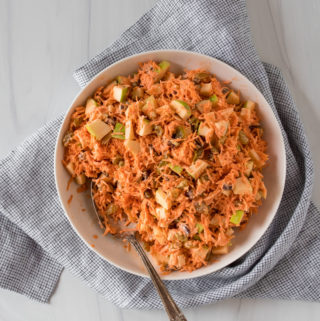 Bright orange carrot apple slaw tossed with apples, raisins, and pecans. Served in a large white bowl on a light striped linen.