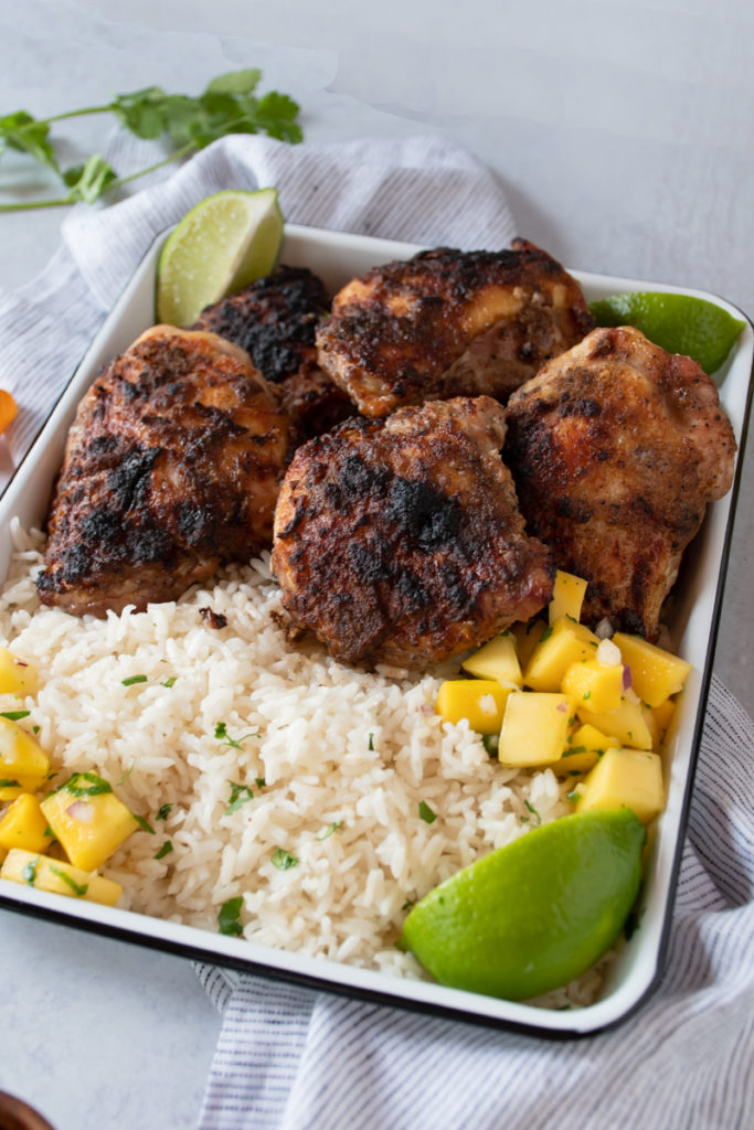 jamaican jerk chicken thighs with blackened crispy skin, smoked on a charcoal grill or in the oven, served on a platter with rice and mango salsa
