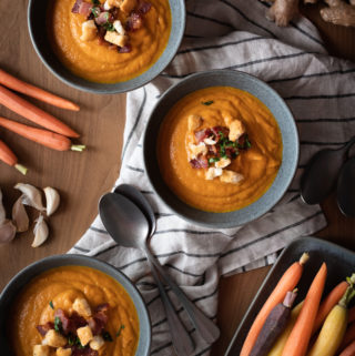 creamy roasted carrot soup served in gray bowls topped with crumbled bacon, chopped basil, and croutons