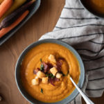 Creamy roasted carrot soup topped with bacon, croutons, and basil served in a gray bowl with carrots and ginger root.