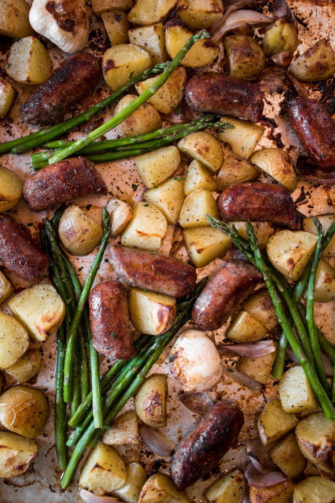 Roasted Italian sausages and crispy potatoes with tender asparagus arranged on a sheet pan with roasted garlic cloves.