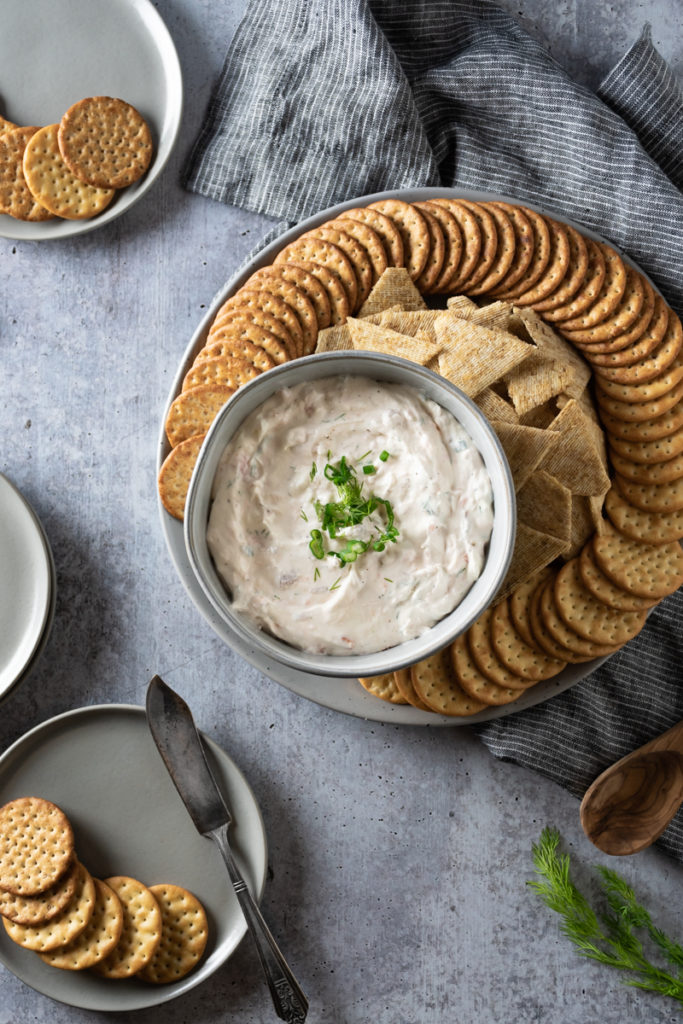 Creamy smoked salmon dip sprinkled with dill and green onions served with a platter of crackers on a gray tray.