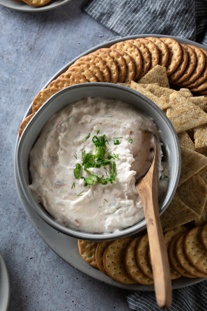 Creamy smoked salmon dip sprinkled with dill and green onions served with a platter of crackers on a gray tray.