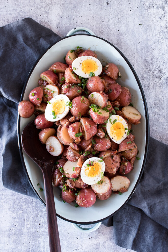 warm potato salad made from red potatoes, served on a blue platter and topped with soft boiled eggs and green onions. 