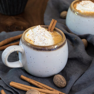Stoneware mug filled to the brim with orangey-hued pumpkin hot chocolate, topped with a dollop of whipped cream and a dusting of nutmeg and a cinnamon stick