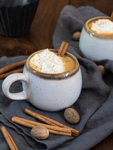Stoneware mug filled to the brim with orangey-hued pumpkin hot chocolate, topped with a dollop of whipped cream and a dusting of nutmeg and a cinnamon stick